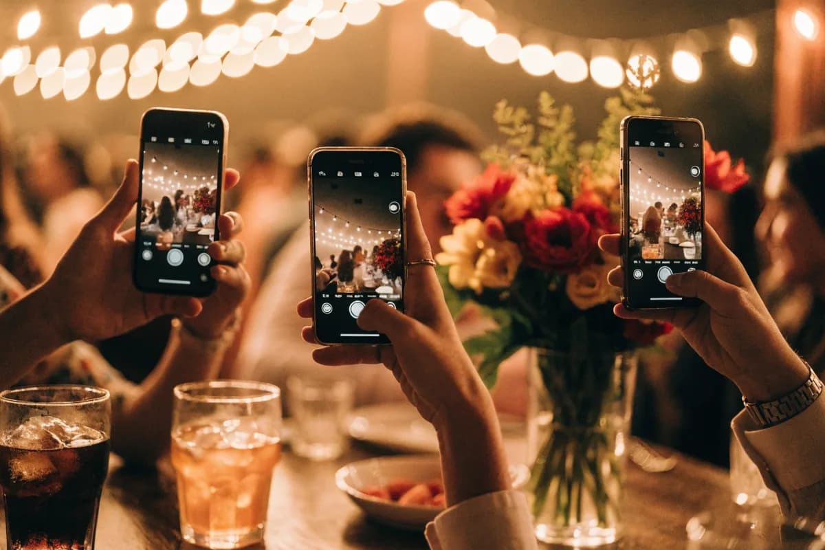 Tres invitados fotografían la fiesta con sus celulares sobre una mesa con flores y luces cálidas de fondo