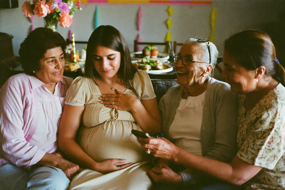 Una mujer embarazada en el sofá de su casa mirando el teléfono junto a su mamá, su abuela y su tía durante un baby shower