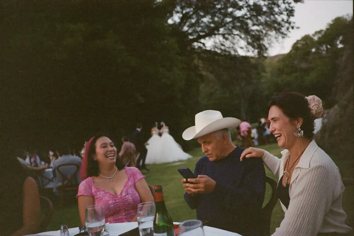 Invitados de una boda al atardecer riendo en su mesa del jardín mientras uno de ellos revisa las fotos en su teléfono y los novios bailan al fondo