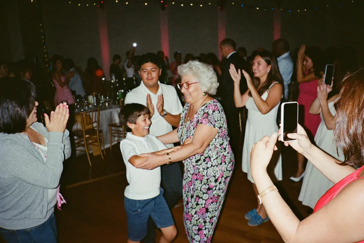 Una abuela bailando con su nieto en la pista de un salón mientras la familia aplaude alrededor y dos parientes graban con sus teléfonos