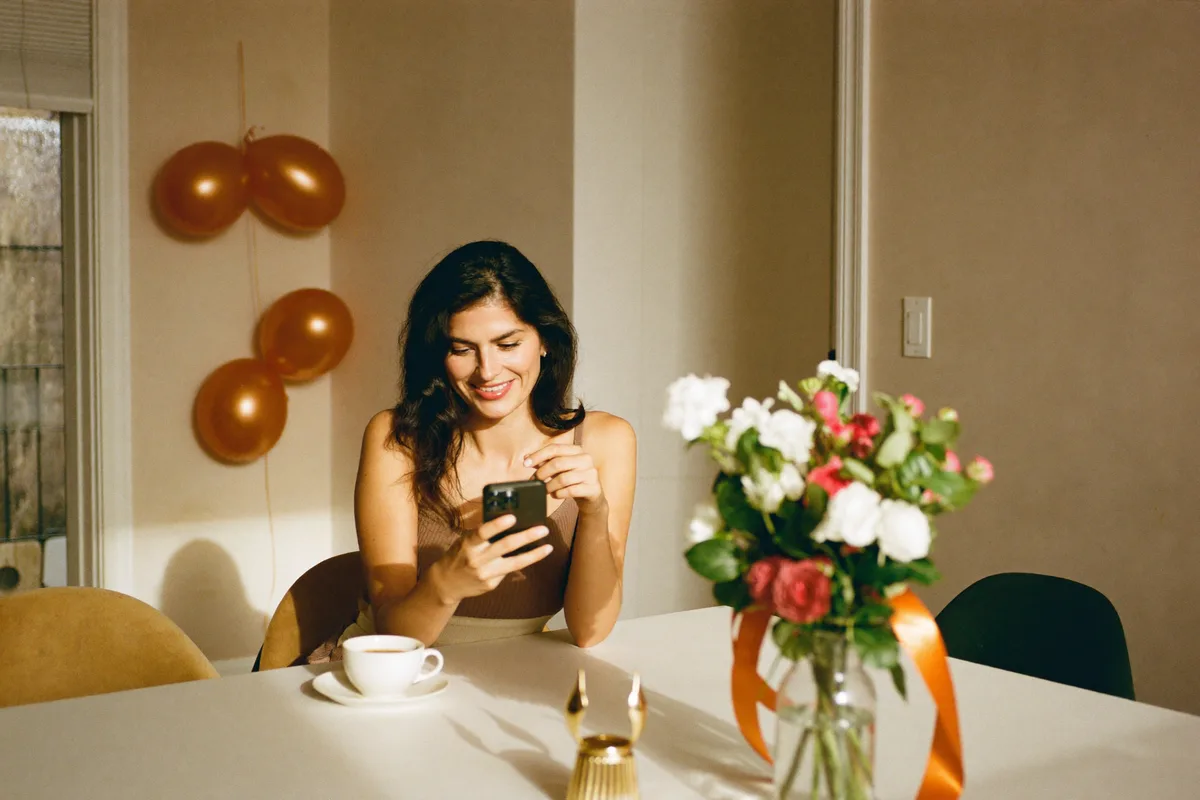 Una mujer en la mesa del comedor, días después de la fiesta, escribe en su celular un mensaje al grupo de la familia para pedir las fotos, con un centro de mesa de la fiesta todavía puesto