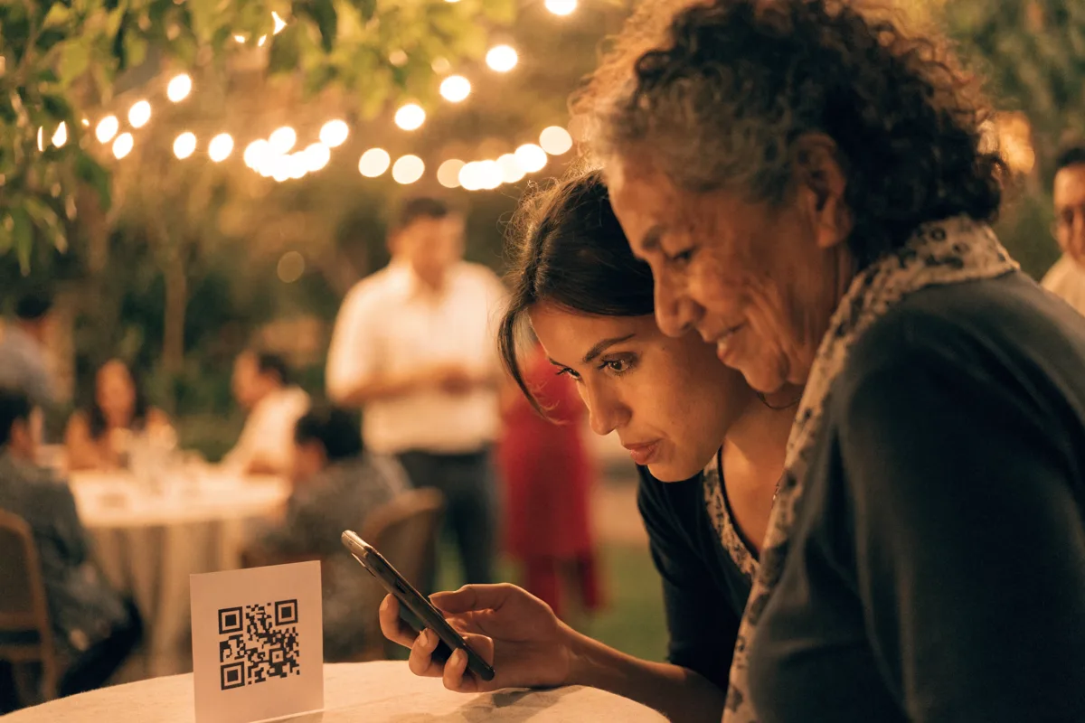 Dos invitadas de distintas generaciones inclinadas juntas sobre el mismo celular en la mesa de una fiesta de jardín de noche: la más joven sostiene el teléfono y la mayor mira la pantalla por encima de su hombro, con la tarjeta del código QR parada sobre el mantel y las series de foquitos y el resto de los invitados desenfocados al fondo