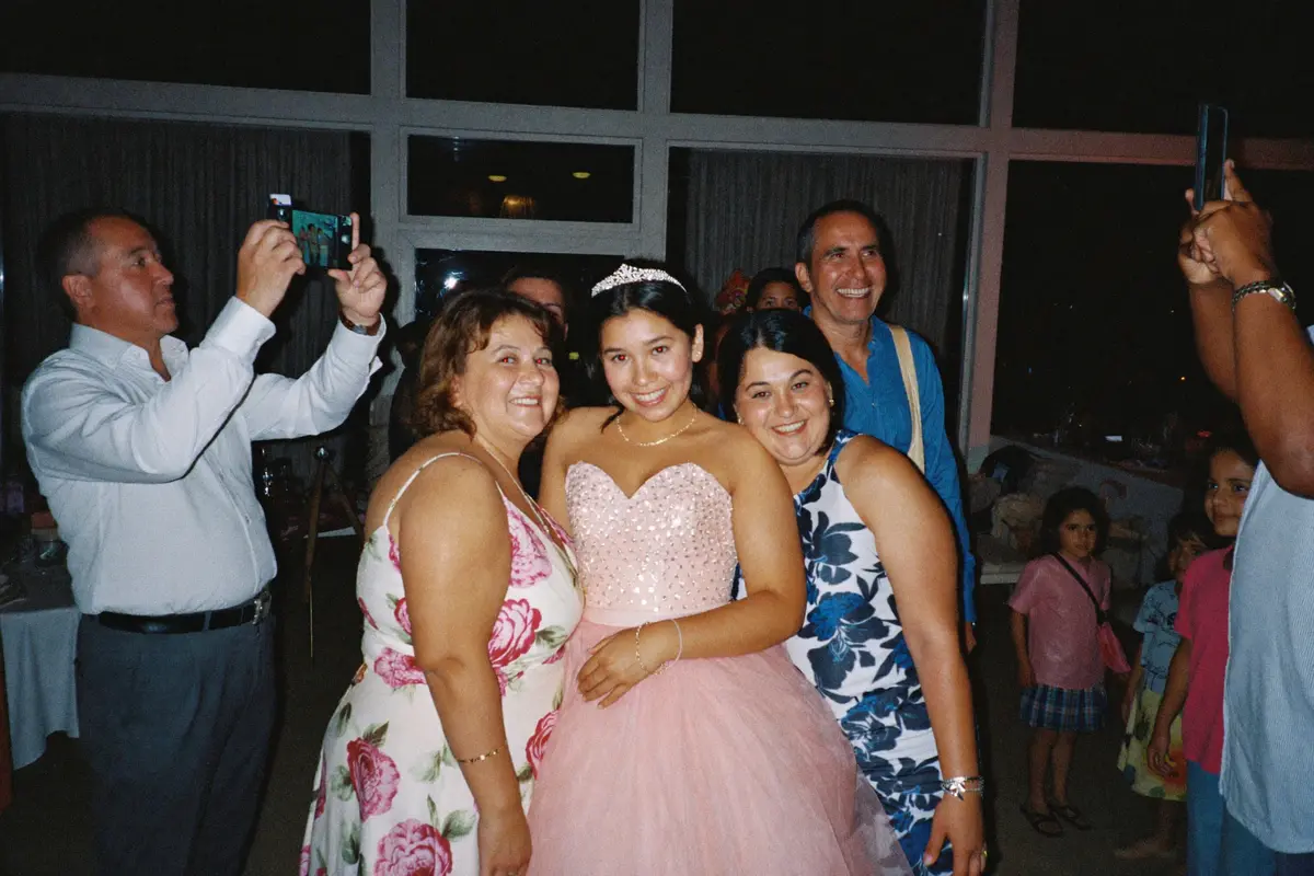 Una quinceañera con su vestido rosa y su tiara, abrazada por su mamá y su tía, mientras dos familiares les toman fotos con el celular en el salón
