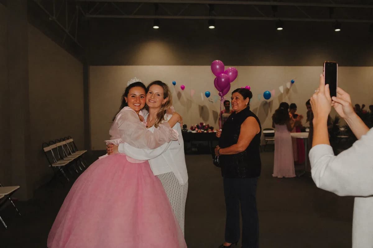 Una quinceañera de vestido rosa y tiara abraza a una invitada mientras alguien les toma una foto con el celular en el salón de la fiesta