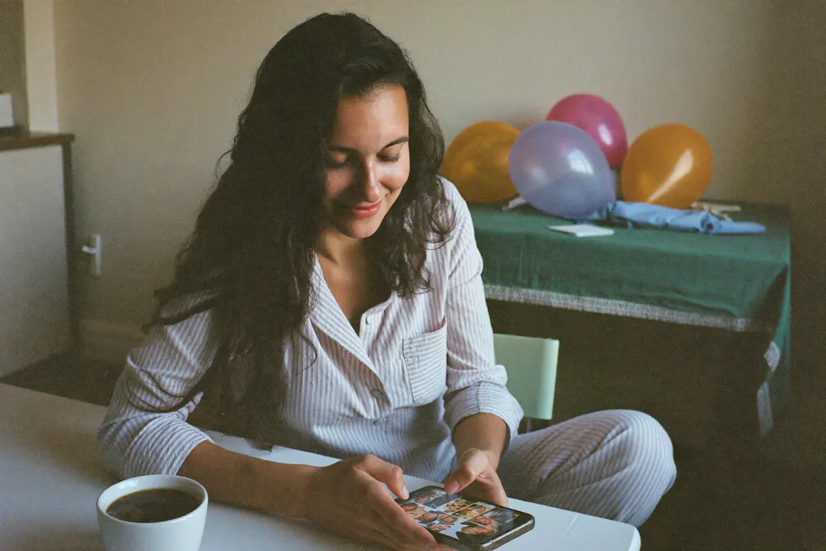 Una mujer en pijama revisa las fotos de la fiesta en su teléfono a la mañana siguiente, con su café y los globos desinflados al fondo