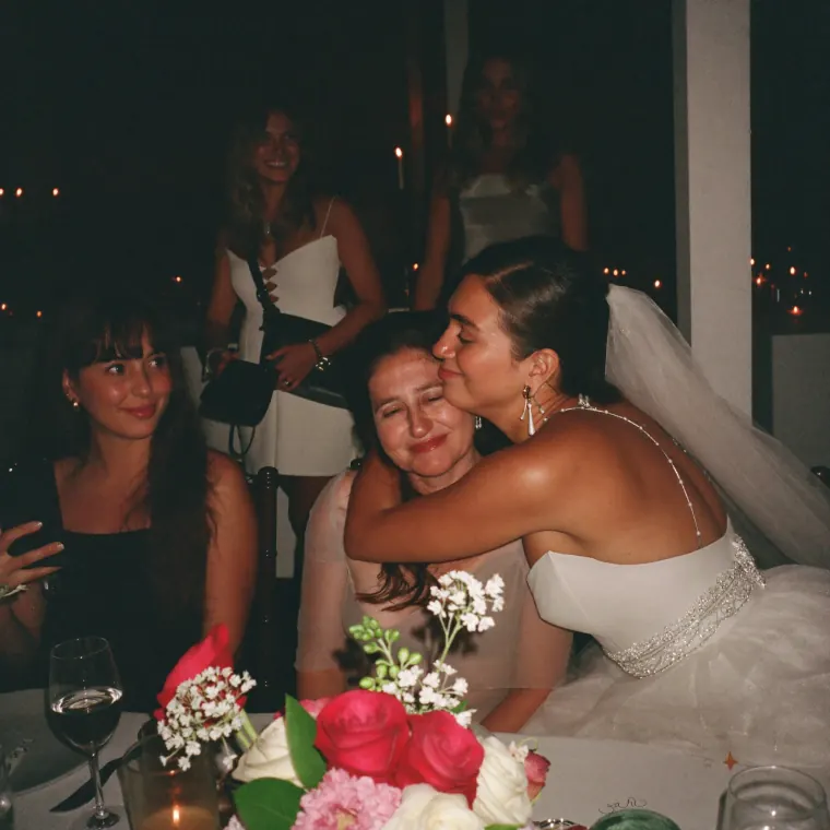 La novia abraza y besa a una mujer de su familia sentada a la mesa, rodeadas de flores y velas.