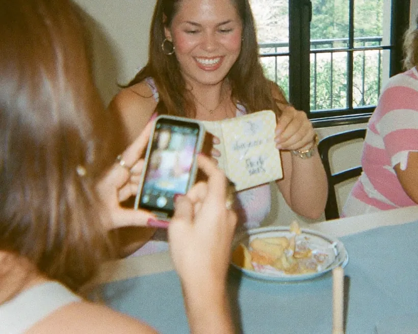 Una invitada fotografía con su celular a la festejada de un baby shower, que sonríe con una tarjeta en la mano junto a la mesa de regalos.