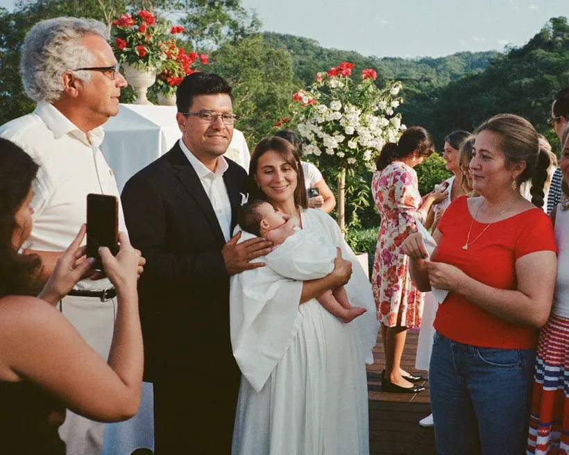 Los papás sostienen a su bebé con el ropón de bautizo en un jardín, rodeados de la familia, mientras una invitada les toma una foto con el celular.