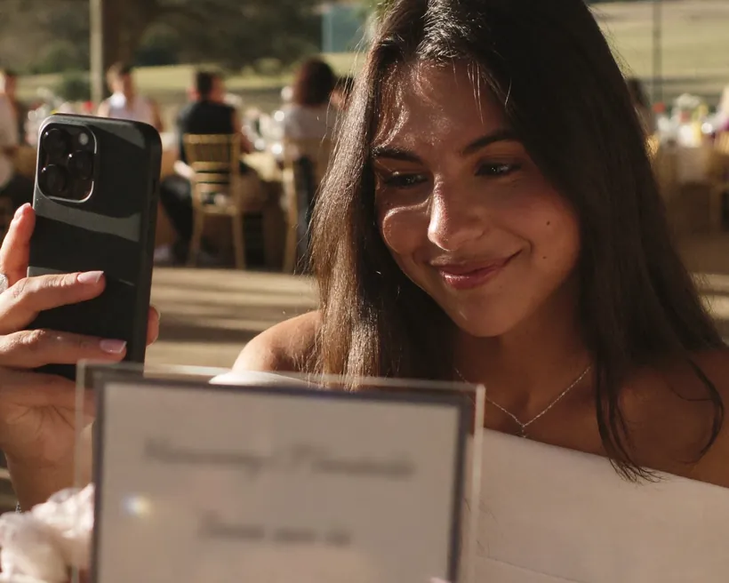 Una invitada de vestido blanco levanta su celular frente a una tarjeta puesta en la mesa de una boda al aire libre, con las mesas del banquete al fondo.